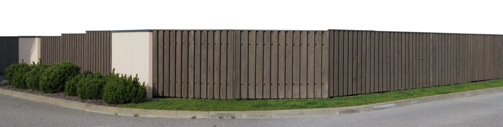 Image of a long, brown-colored security fence made out of boards protecting a business establishment. The fence wraps around a street corner and there is some green shrubbery in front of a portion of the fence. 